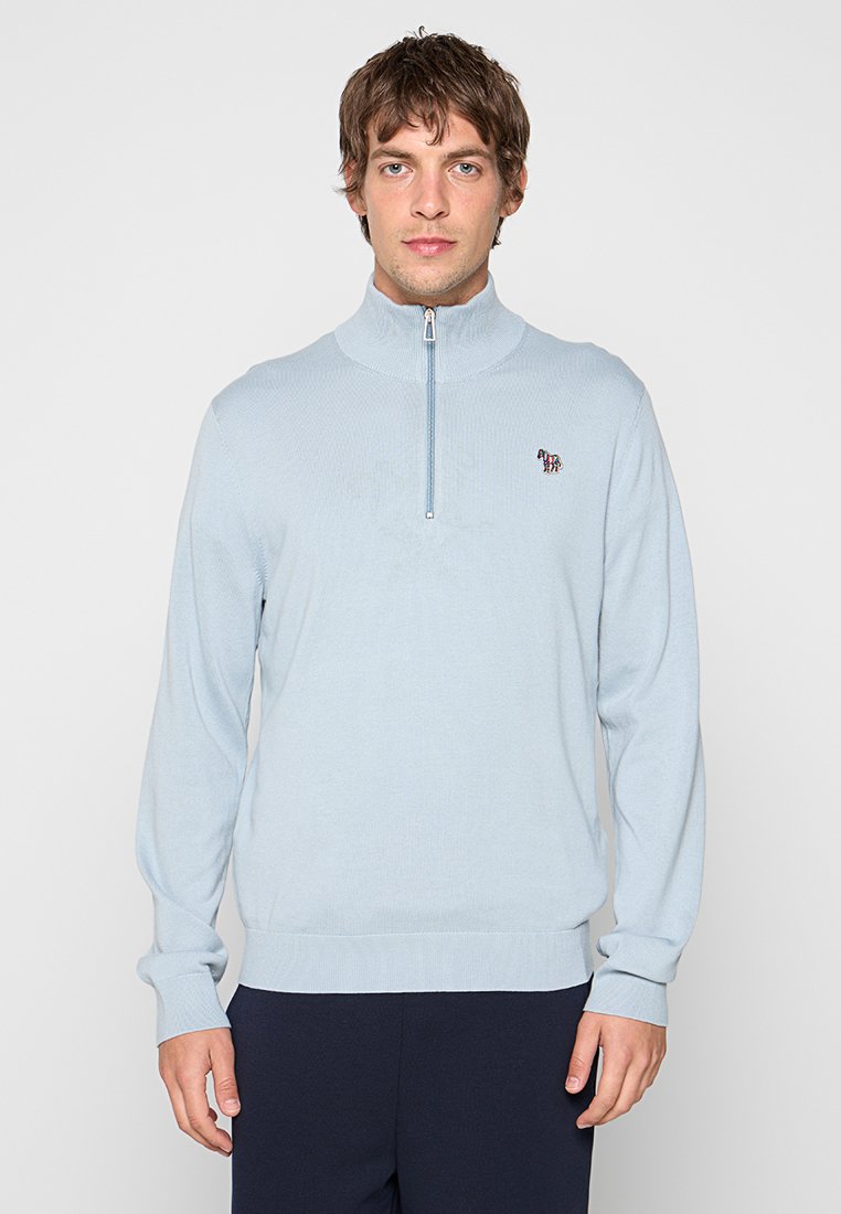 Mann trägt hellblauen Viertel-Reißverschluss-Pullover mit kleinem gesticktem Zebra auf der Brust und steht vor einfachem hellem Hintergrund.
