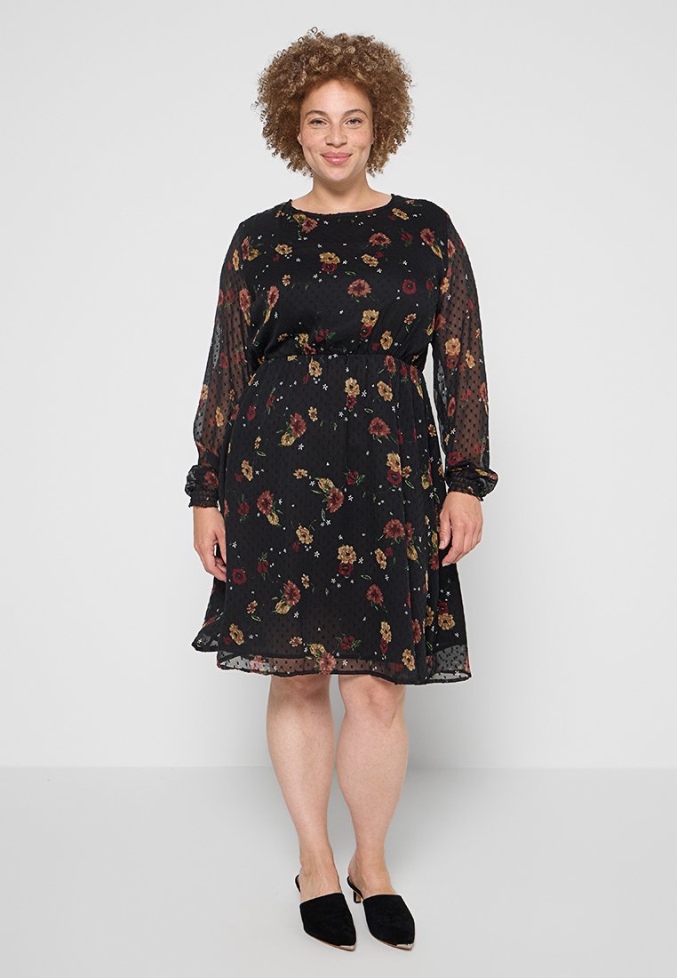 Schwarzes Kleid mit Blumendruck, transparenten langen Ärmeln, elastischem Bund, fließendem Rock und strukturiertem Stoff. Getragen mit schwarzen Slippern.