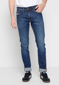 Ein Mann trägt hochgekrempelte dunkelblaue Slim-Fit-Jeans und schwarze Adidas-Sneaker mit weißen Streifen, während er vor einem schlichten hellen Hintergrund steht.