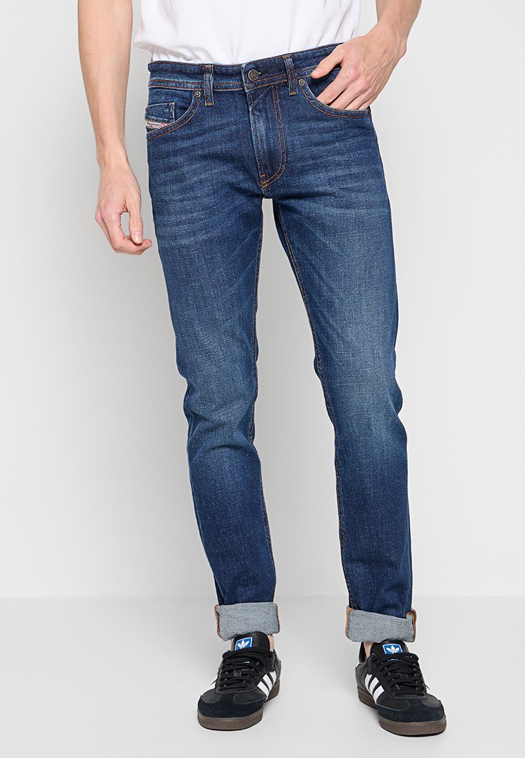 Ein Mann trägt hochgekrempelte dunkelblaue Slim-Fit-Jeans und schwarze Adidas-Sneaker mit weißen Streifen, während er vor einem schlichten hellen Hintergrund steht.