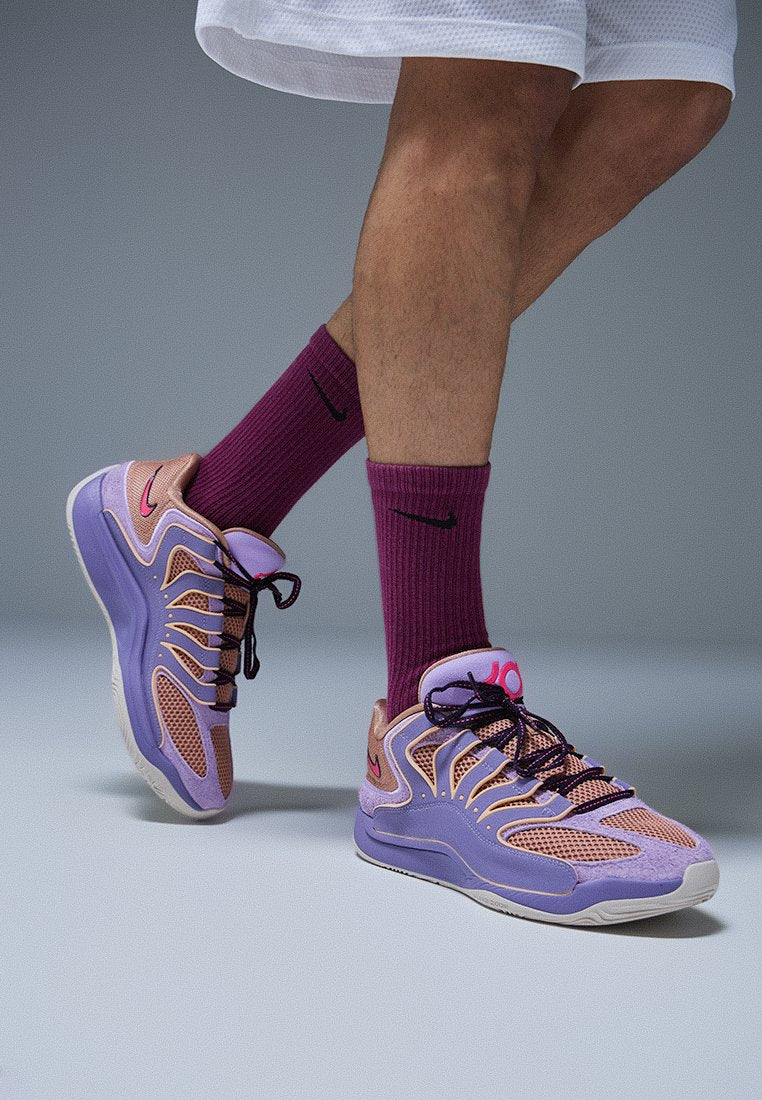 Person trägt lila und pfirsichfarbene Sportschuhe mit passenden weinroten Nike-Socken und weißen Shorts, steht vor grauem Hintergrund.