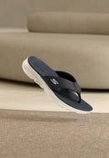 Marineblaue Skechers Flip-Flops mit weißer Sohle, die über einem beigen Teppich schweben, im Hintergrund unscharfes beiges Sofa.