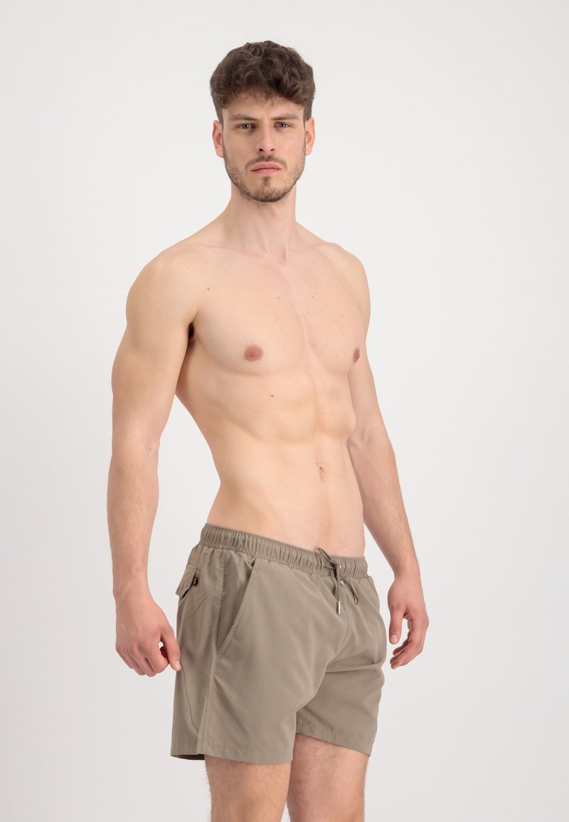 Hemdloser Mann mit kurzem braunem Haar, der beige Badeshorts trägt, steht vor einem einfarbigen hellen Hintergrund, schaut nach vorne und hat die Arme locker.