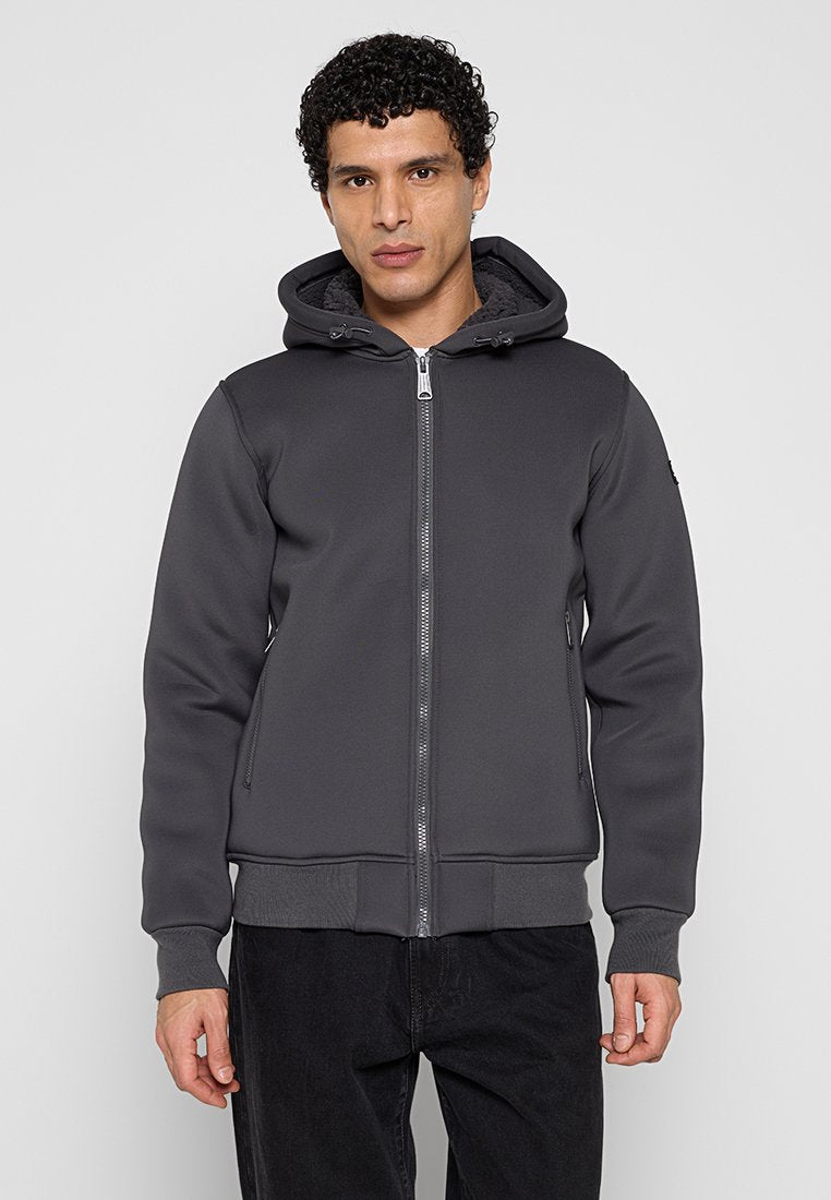 Mann trägt eine dunkelgraue Kapuzenjacke mit Reißverschluss und Taschen, steht vor einem schlichten hellgrauen Hintergrund.