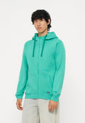 Junger Mann mit lockigem Haar, der einen türkisfarbenen Hoodie mit Reißverschluss und hellgraue Hose trägt, steht vor einem schlichten weißen Hintergrund.