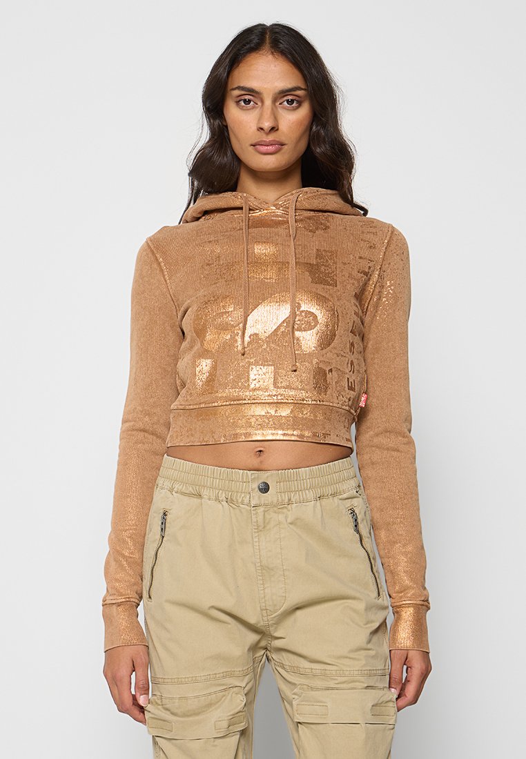 Zuscchnittenes Hoodie in einer metallischen kupfernen Farbe mit einer strukturierten Oberfläche und Logo. Kombiniert mit beigen Cargo-Hosen mit Reißverschlusstaschen.