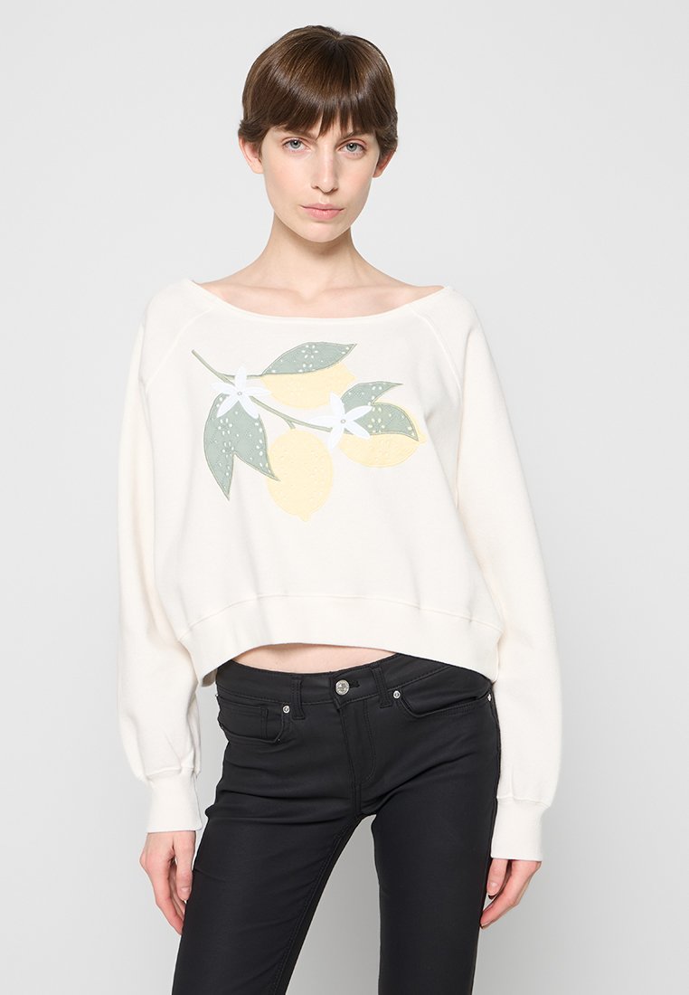 Junge Frau trägt einen weißen Pullover mit Zitronen- und Blättergrafik, kombiniert mit schwarzen, enganliegenden Jeans, steht vor einem schlichten Hintergrund.
