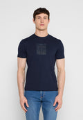 Mann trägt ein marineblaues, eng anliegendes T-Shirt mit dezentem geometrischem Logo und blaue Jeans, steht vor einem schlichten hellgrauen Hintergrund.