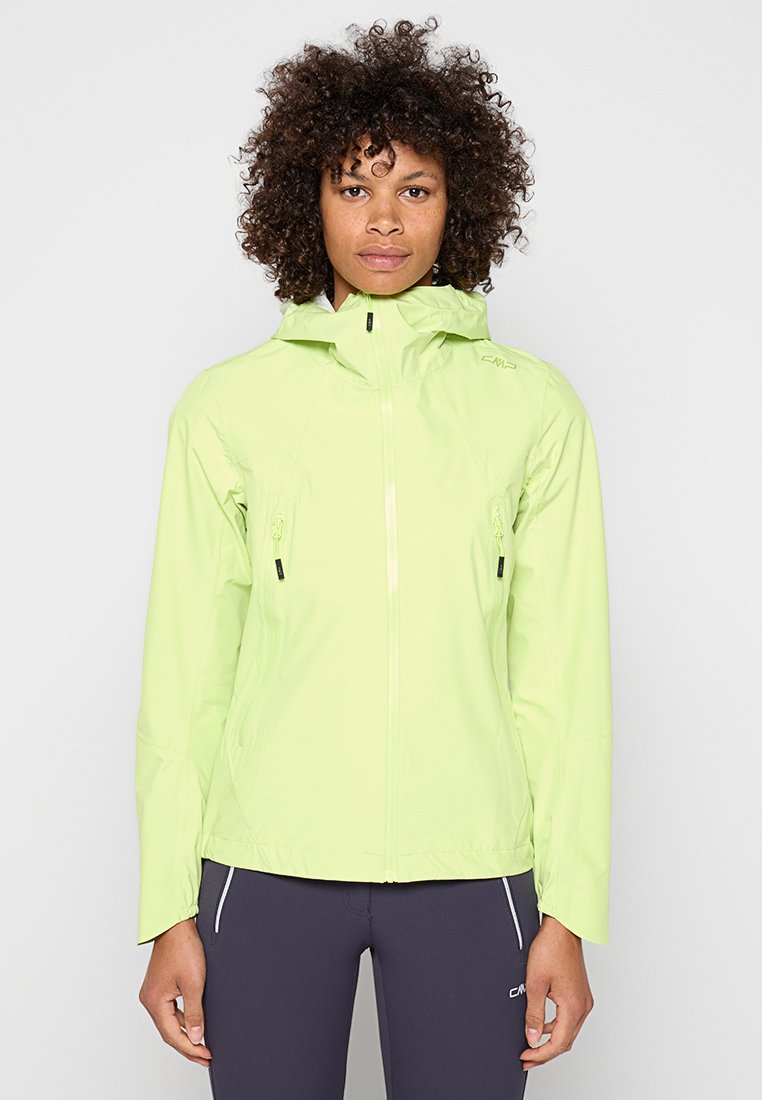Frau mit lockigem Haar, die eine hellgrüne Kapuzenjacke und dunkelgraue Sporthose trägt, steht mit dem Gesicht nach vorne vor einem schlichten Hintergrund.