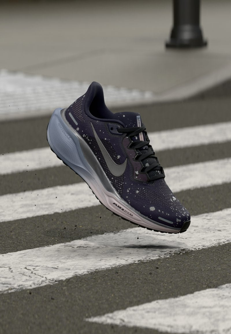 Schwarzer und grauer Nike Air Zoom Laufschuh mit weißen Sprenkeln, der in der Luft über einem Fußgängerüberweg auf Asphalt angezeigt wird.