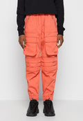 Person, der korallenfarbene Utility-Hosen mit mehreren Taschen und schwarze Sneakers trägt, steht vor einem einfarbigen hellen Hintergrund.