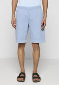 Mann in hellblauen Shorts mit elastischem Bund, einem weißen Hemd und schwarzen Sandalen mit Schnalle, der vor einem schlichten Hintergrund steht.
