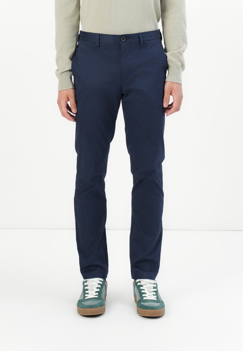 Marineblaue Chinos aus leichtem Material; mit schmaler Passform, Gürtelschlaufen, frontalen Taschen und Knopfverschluss. Kombiniert mit grünen Sneakern.