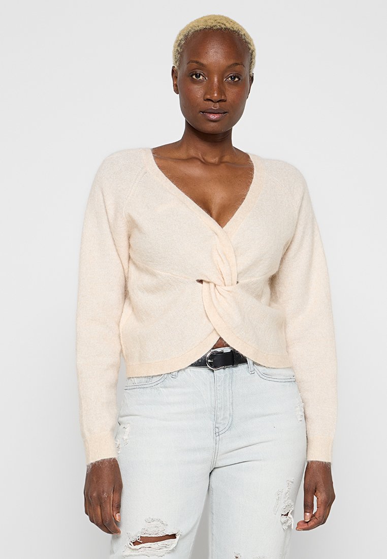 Hellbeige gestrickte Strickjacke mit V-Ausschnitt und verdrehtem Vorderdetail. Verfügt über lange Ärmel und eine weiche Textur, kombiniert mit hellen, zerrissenen Jeans.