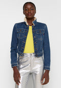 Jeansjacke mit Schafsfellkragen, mit Knopfleiste, Brusttaschen und tailliertem Schnitt. Getragen über einem gelben Oberteil und silbernen Hosen.