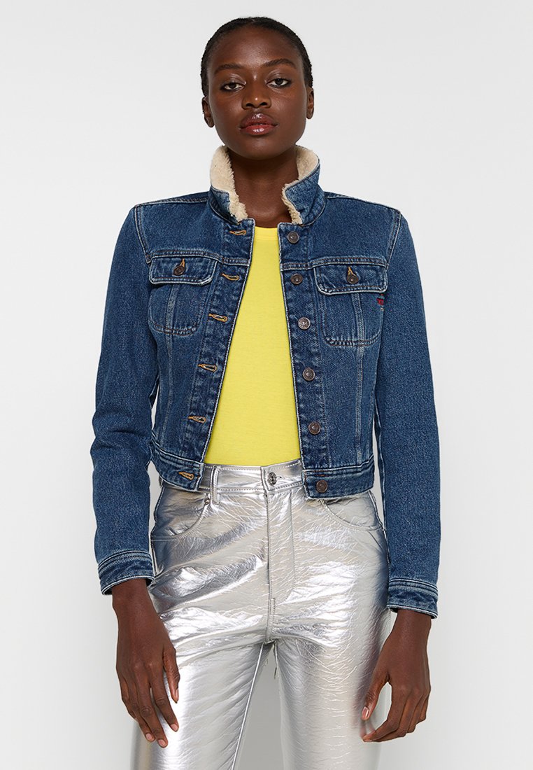 Jeansjacke mit Schafsfellkragen, mit Knopfleiste, Brusttaschen und tailliertem Schnitt. Getragen über einem gelben Oberteil und silbernen Hosen.