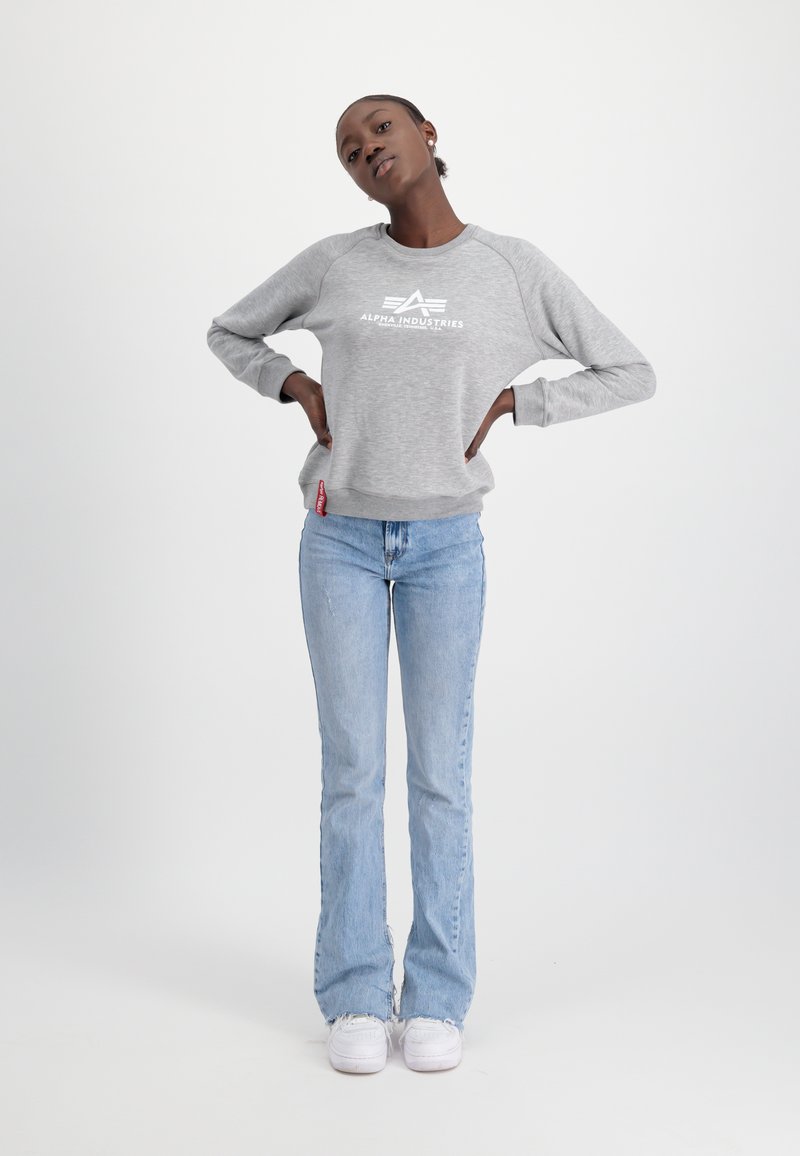 Grau sweatshirt mit weißem "Alpha Industries"-Druck, kombiniert mit hellblauen Schlagjeans und weißen Sneakern vor einem einfarbigen Hintergrund.