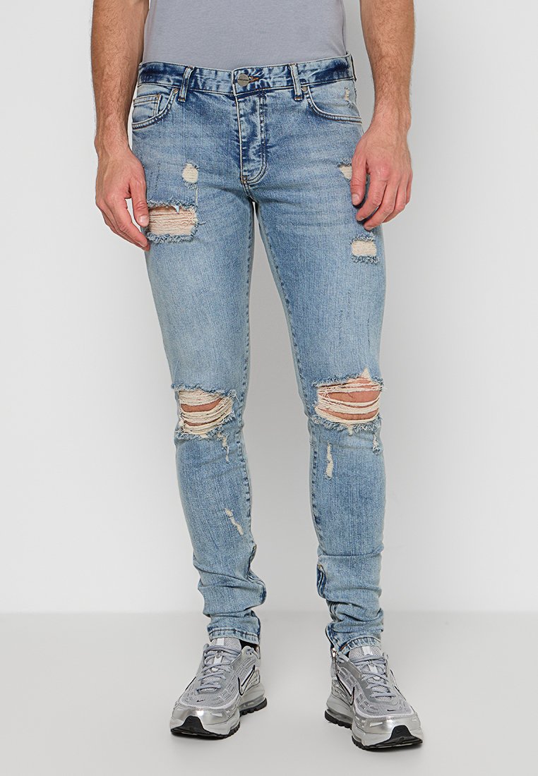 Helle blaue, schmale Jeans mit ausgefransten Löchern an den Knien und Oberschenkeln, aus Denim. Getragen mit grauen Sportschuhen.
