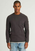 Dunkelgrauer Strickpullover mit Rundhalsausschnitt, langen Ärmeln, Fronttasche und schwarzem Logoakzent. Getragen über hellblauen Jeans.