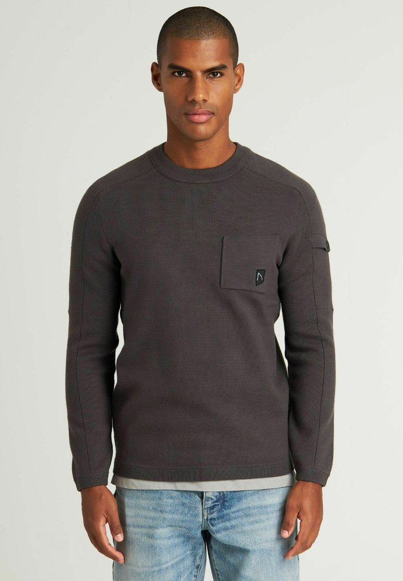 Dunkelgrauer Strickpullover mit Rundhalsausschnitt, langen Ärmeln, Fronttasche und schwarzem Logoakzent. Getragen über hellblauen Jeans.