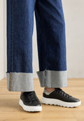 Schwarze Sneakers mit strukturiertem Obermaterial, weißen Schaumsohlen, kombiniert mit blauen Jeans-Hosen im weiten Schnitt mit ausgefranstem Saum und kontrastierenden grauen Bündchen.