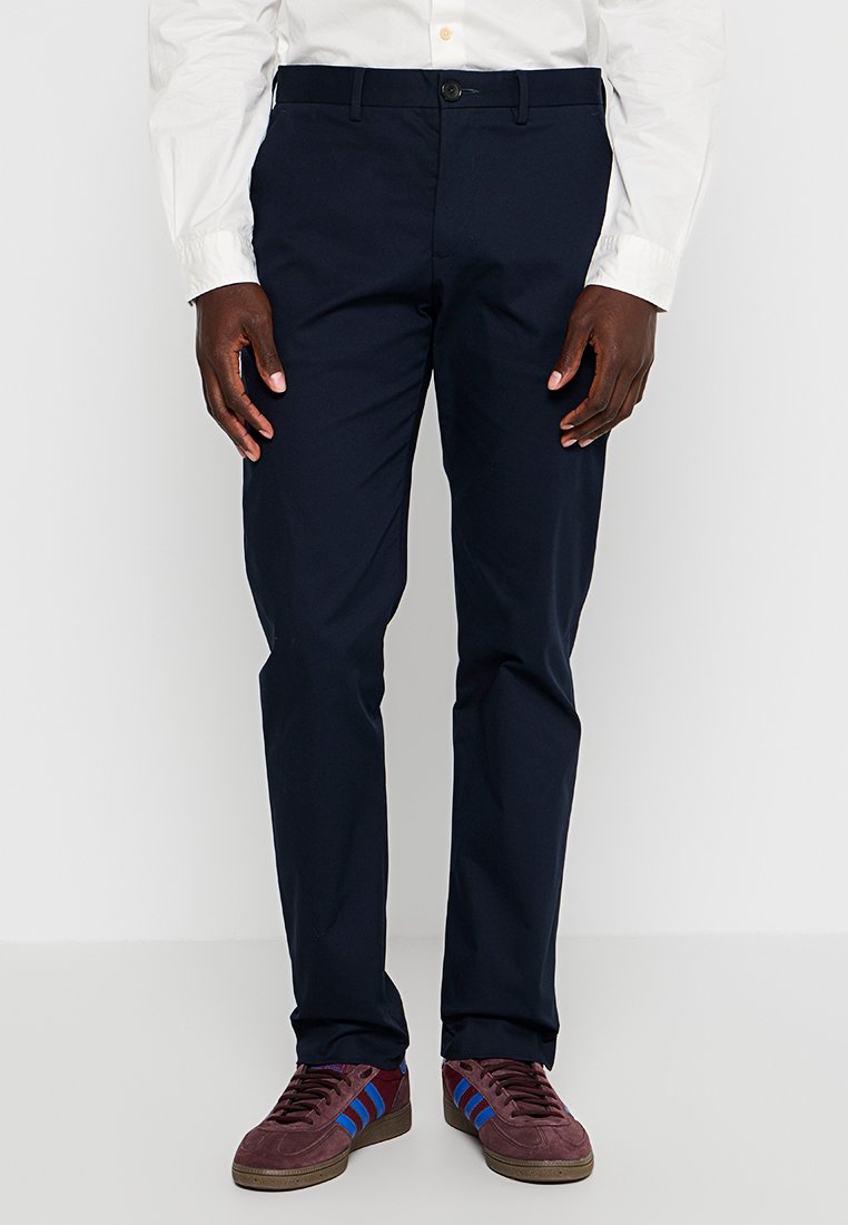 Navyblaue Hose aus glattem Stoff, mit klassischer Passform und einem einzelnen Knopfverschluss vorne. Kombiniert mit bordeauxfarbenen Sneakers mit blauen Streifen.