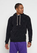Mann mit geflochtenem Haar, der einen schwarzen Kapuzenpullover mit Muster und lila Shorts trägt, schaut nach rechts vor einem schlichten weißen Hintergrund.
