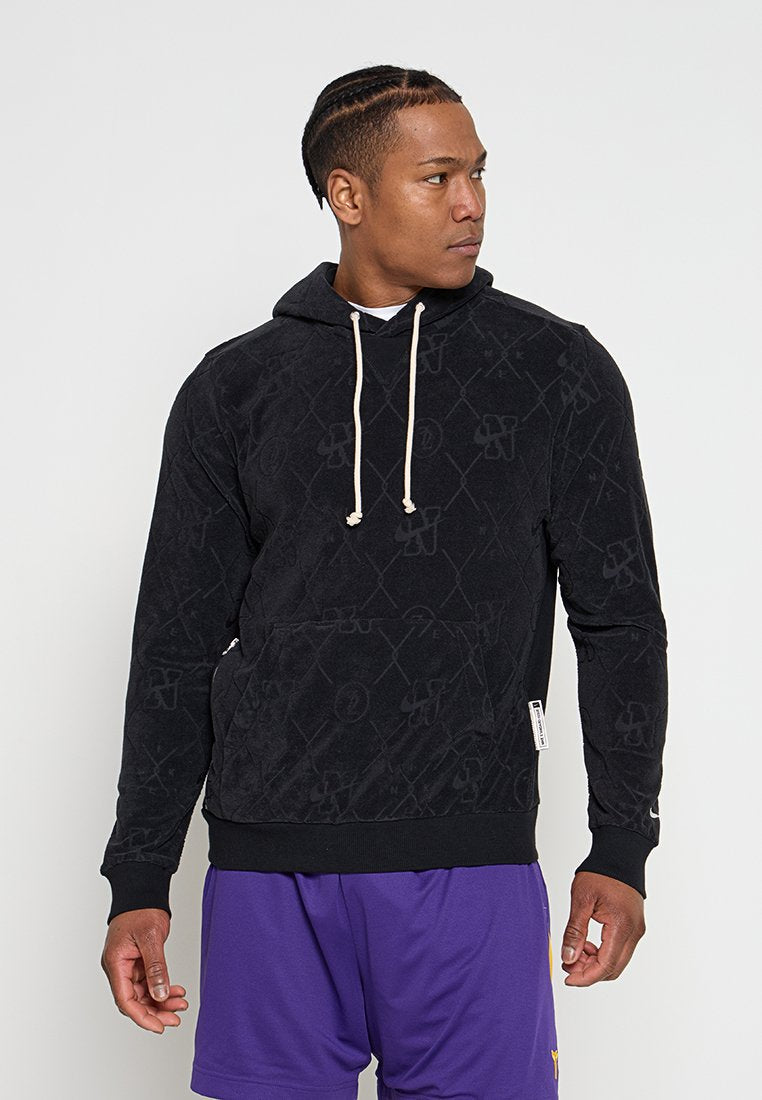 Mann mit geflochtenem Haar, der einen schwarzen Kapuzenpullover mit Muster und lila Shorts trägt, schaut nach rechts vor einem schlichten weißen Hintergrund.