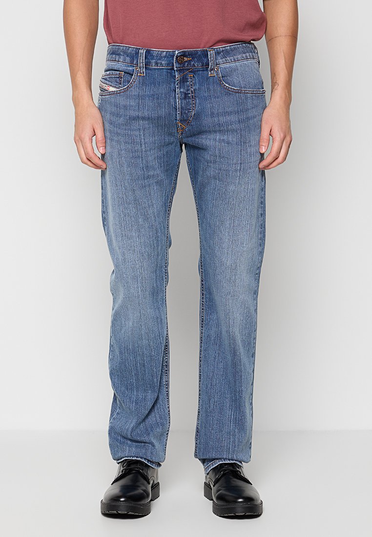 Blaue Denim-Jeans mit geradem Bein-Design, fünf Taschen, kontrastierenden Nähten und subtilen Verwaschungen. Getragen mit schwarzen Schuhen.