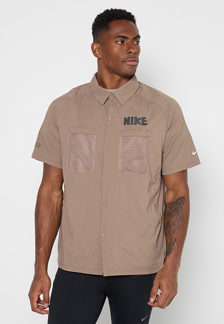 Mann mit kurzen, lockigen Haaren, der ein beiges, kurzärmliges Hemd mit zwei Netz-Taschen und schwarzem Nike-Logo auf Brust und Ärmel trägt.