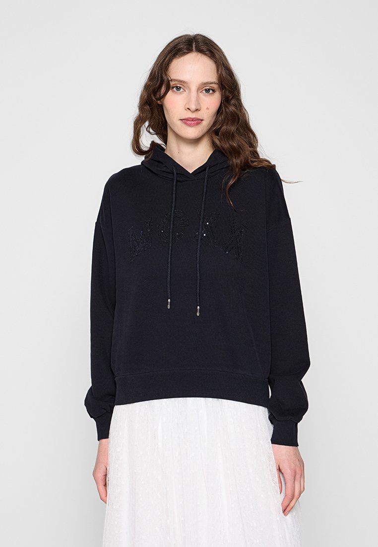 Frau mit langen, welligen Haaren, die einen schwarzen Max-Hoodie mit Kordelzügen und einen weißen Rock trägt, stehend vor einem schlichten, hellen Hintergrund.