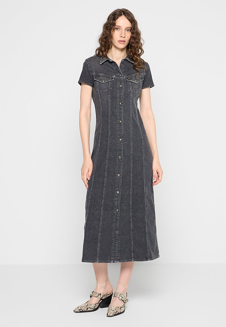 Frau mit lockigem Haar, die ein schwarzes knöpfen Midi-Kleid aus Denim mit kurzen Ärmeln und Schlangenledermuster-Absatzschuhen trägt und steht.