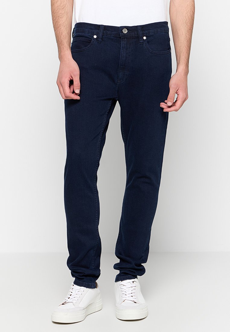 Marineblaue Jeans aus Denim mit schmaler Passform, sichtbaren Nähten, Five-Pocket-Design und Knopfverschluss. Getragen mit einem schlichten weißen T-Shirt und Sneakers.