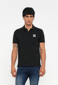 Schwarzes Poloshirt aus Baumwolle mit kurzem Kragen, zwei Knöpfen und einem kleinen weißen quadratischen Logo auf der Brust. Tragen mit blauen Jeans.