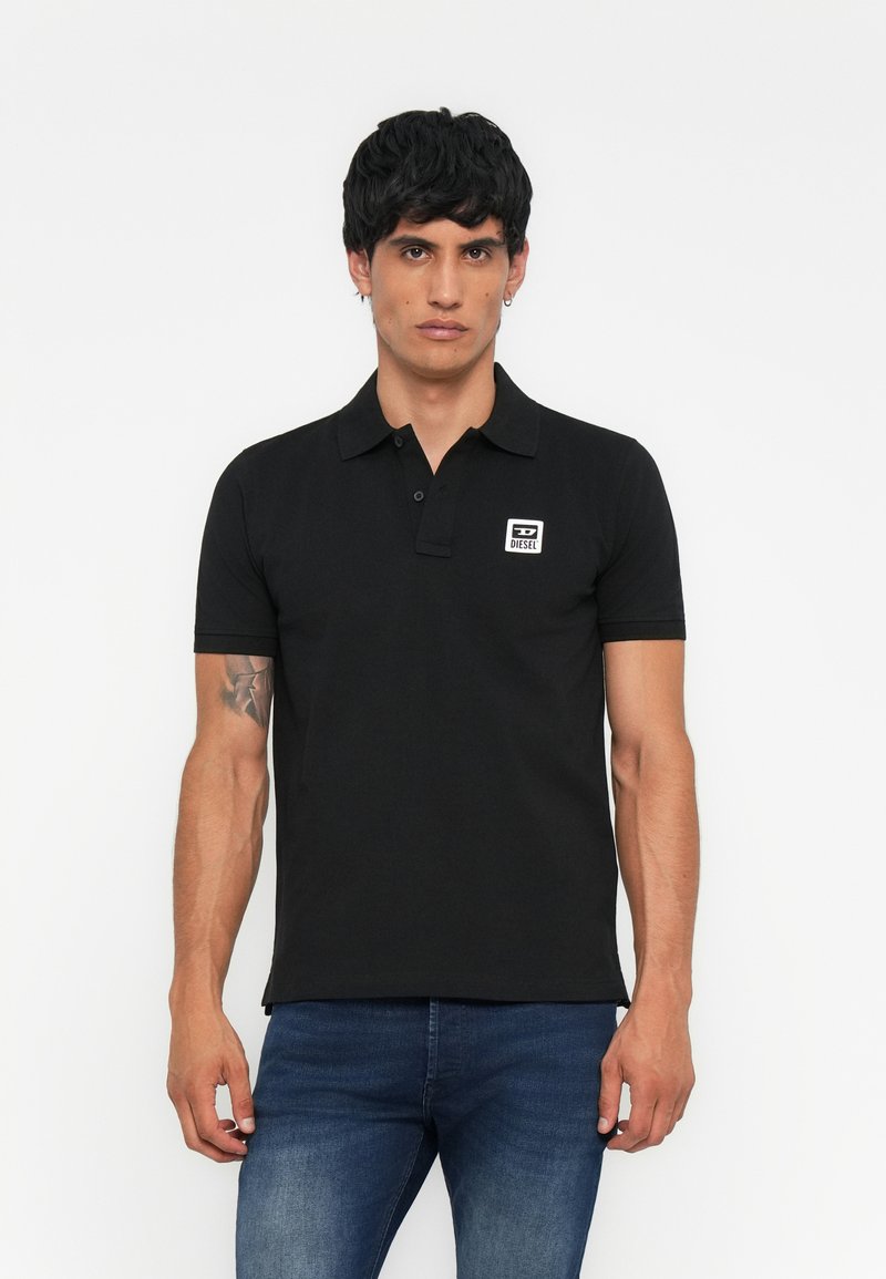 Schwarzes Poloshirt aus Baumwolle mit kurzem Kragen, zwei Knöpfen und einem kleinen weißen quadratischen Logo auf der Brust. Tragen mit blauen Jeans.