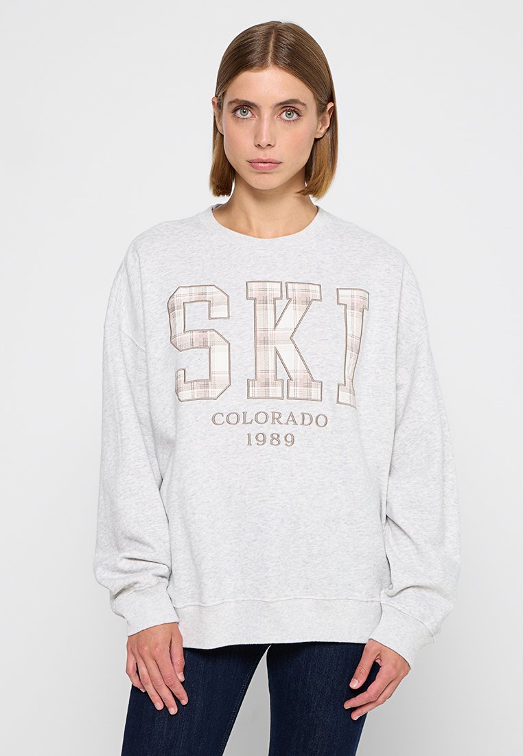 Grauer Sweatshirt mit langen Ärmeln, das in der Mitte das Wort "SKI" in fettem Karomuster und "COLORADO 1989" in braunem Text zeigt. Weiches Material.