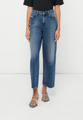 Frau in hochgeschnittenen, geraden blauen Jeans mit ausgewaschenen Details und schwarzen Riemchen-Absatzsandalen vor einem weißen Hintergrund.