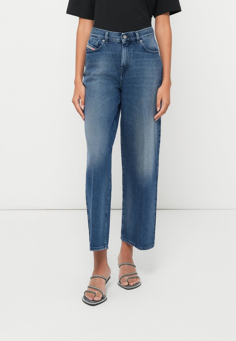 Frau in hochgeschnittenen, geraden blauen Jeans mit ausgewaschenen Details und schwarzen Riemchen-Absatzsandalen vor einem weißen Hintergrund.
