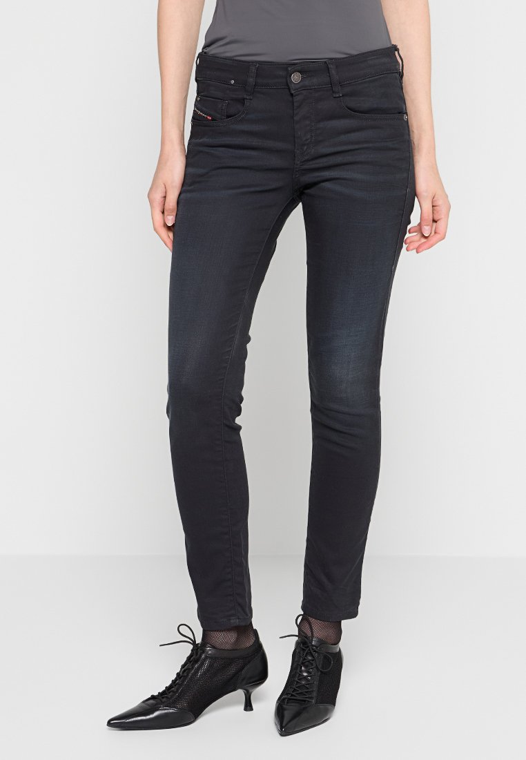 Person in schmalen schwarzen Jeans, schwarzen spitzen Schnürschuhen mit Kitten-Heels und einem grauen Oberteil, stehend vor einem schlichten Hintergrund.