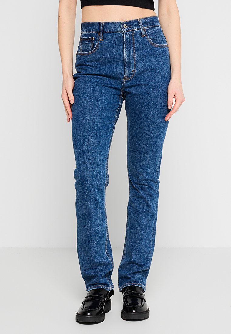 Person trägt hoch taillierte, blaue gerade Jeans und schwarze klobige Loafer, ein kurzes schwarzes Oberteil ist über der Taille sichtbar.