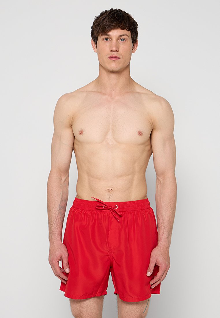 Fit junger Mann steht, trägt leuchtend rote Badeshorts mit Kordelzug, oben ohne vor einem einfarbigen hellgrauen Hintergrund.