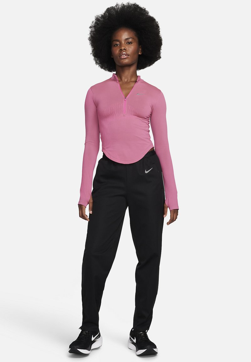 Frau mit natürlichem Afro-Haarstil, die ein rosa langärmliges Zip-Top, schwarze Nike Jogginghosen und schwarze Nike Sneakers trägt, steht frontally.