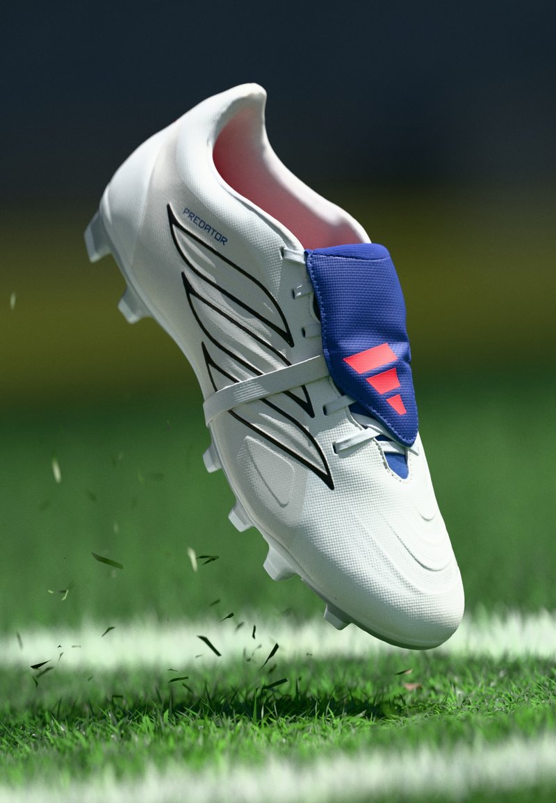 Weißer Fußballschuh mit strukturierter Oberseite, blauem Akzent, schwarzen Streifen und pinkem Logo. Der Schuh ist teilweise vom Gras abgehoben, wodurch die Stollen sichtbar sind.