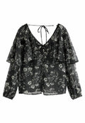Schwarze Bluse mit floralem Muster, V-Ausschnitt, Rüscheln an den Ärmeln und einer Schleife im Rücken. Hat weiße Blumenmuster auf dem durchscheinenden Stoff.