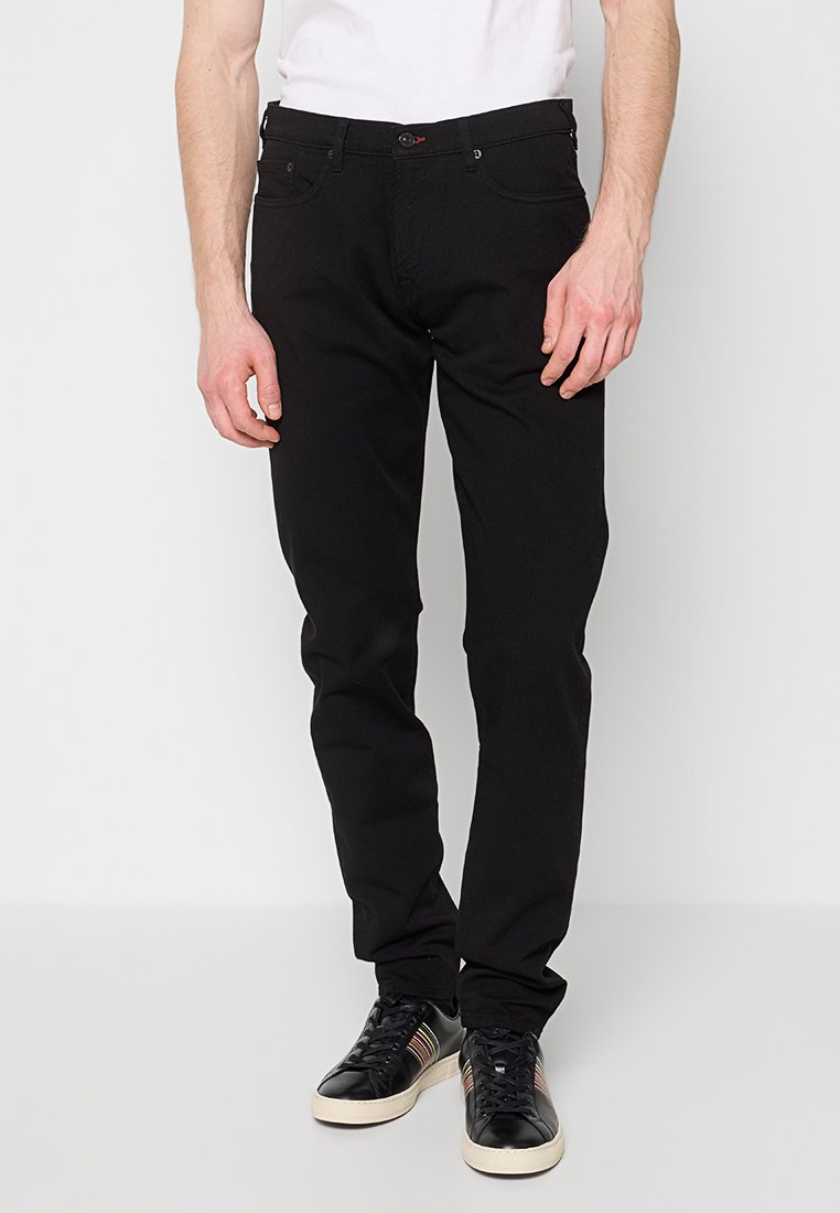 Schwarze Jeans mit geradem Bein, fünf Taschen und dezenten Nähten. Gekleidet mit weißem T-Shirt und schwarzen Turnschuhen.