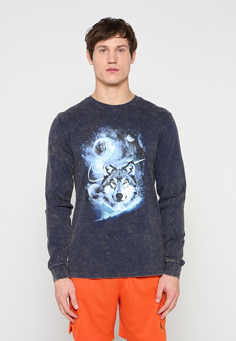 Junger Mann trägt ein verblasstes schwarzes Langarmshirt mit einer blauen Wolfsgrafik und leuchtend orangefarbene Sportshorts.
