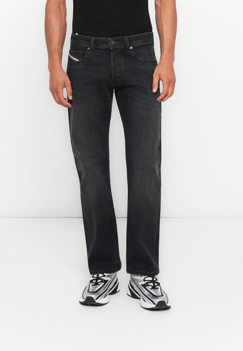 Mann trägt schwarze gerade Jeans mit Vorder- und Gesäßtaschen, kombiniert mit silber-schwarzen Sportschuhen, stehend vor einem weißen Hintergrund.