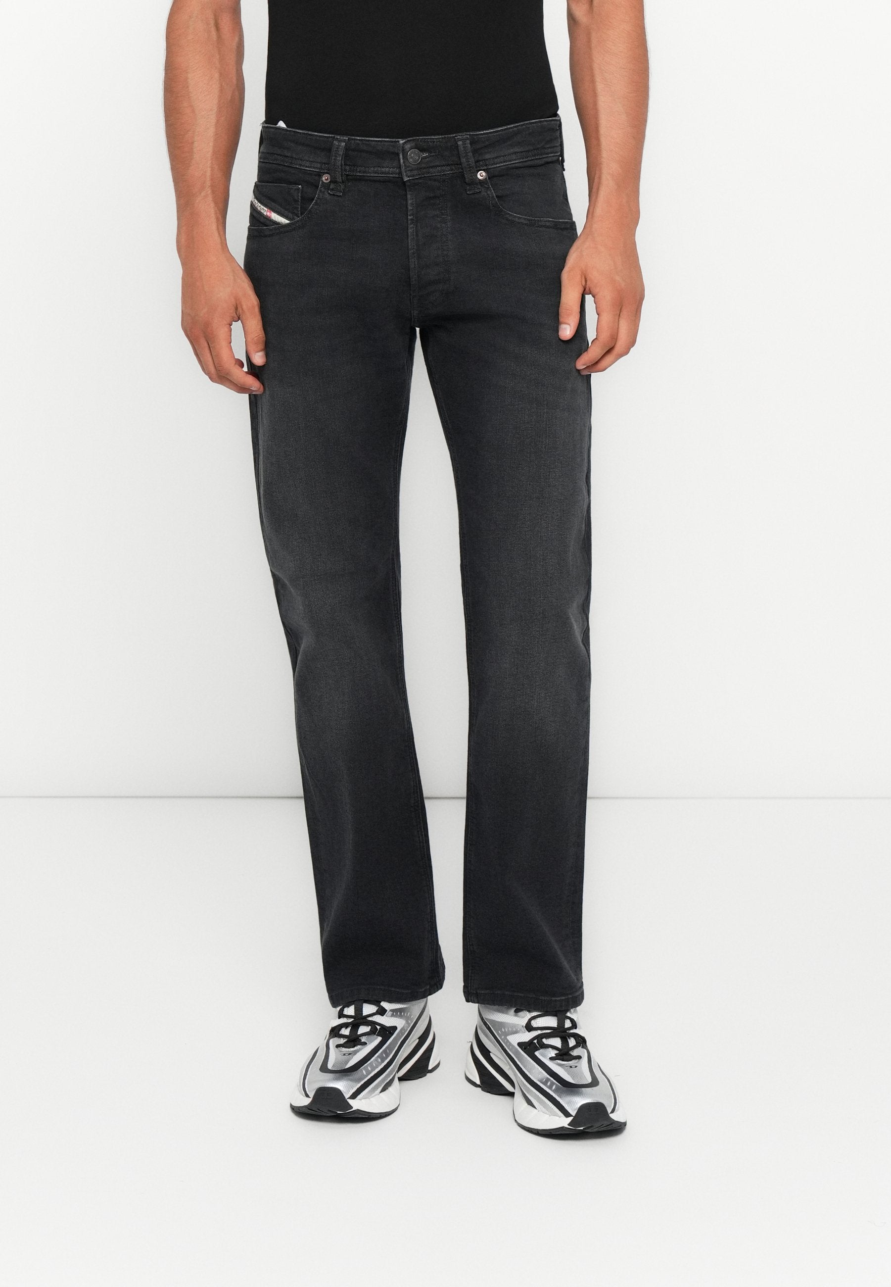 Mann trägt schwarze gerade Jeans mit Vorder- und Gesäßtaschen, kombiniert mit silber-schwarzen Sportschuhen, stehend vor einem weißen Hintergrund.