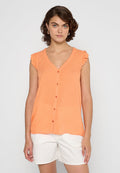 Frau mit kurzem lockigem Haar, die ein oranges ärmelloses Oberteil mit Knopfleiste und Rüschen an den Schultern sowie eine weiße bestickte Shorts trägt.