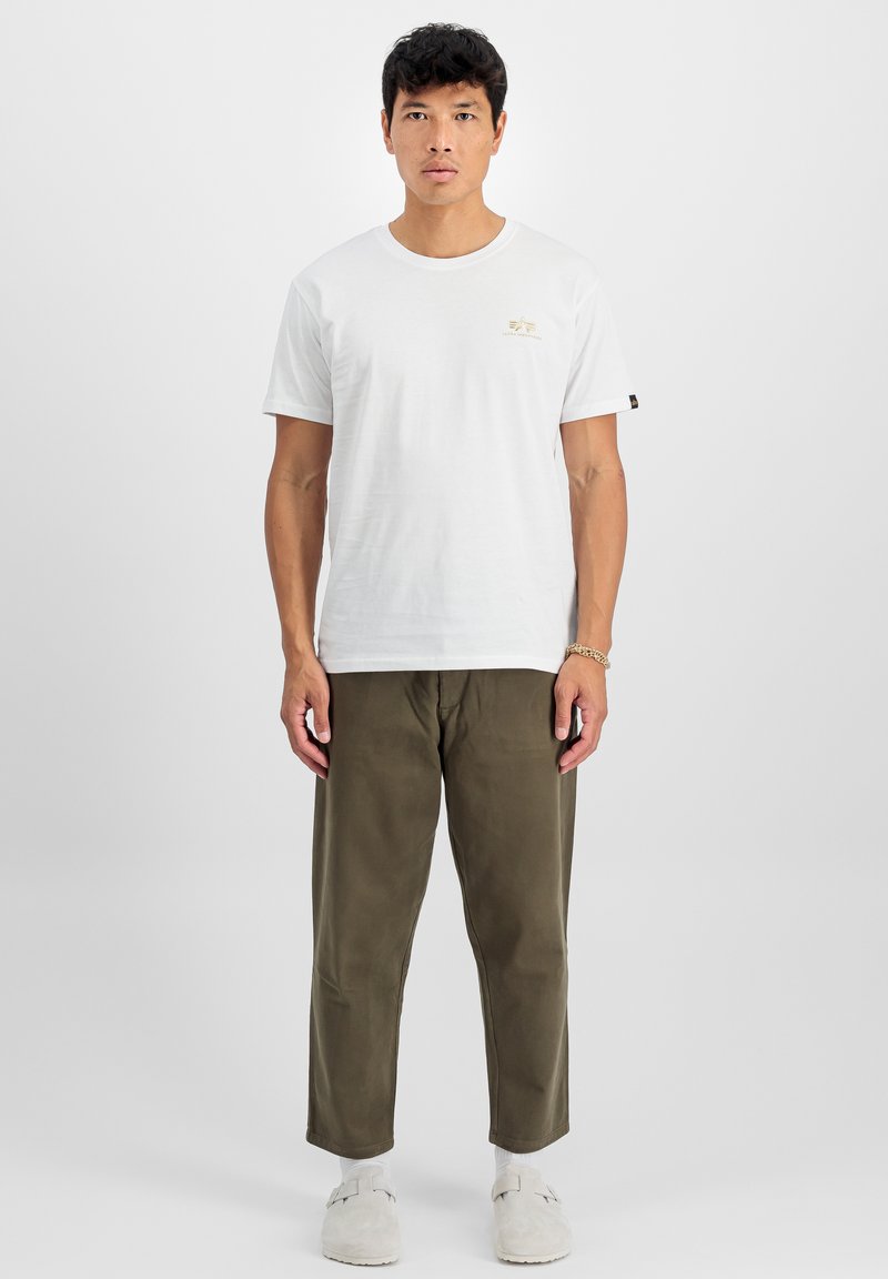 Weißes Baumwoll-T-Shirt mit Rundhalsausschnitt und kleinem goldenen Logodetail, kombiniert mit olivgrünen Hosen und hellgrauen Slippern.